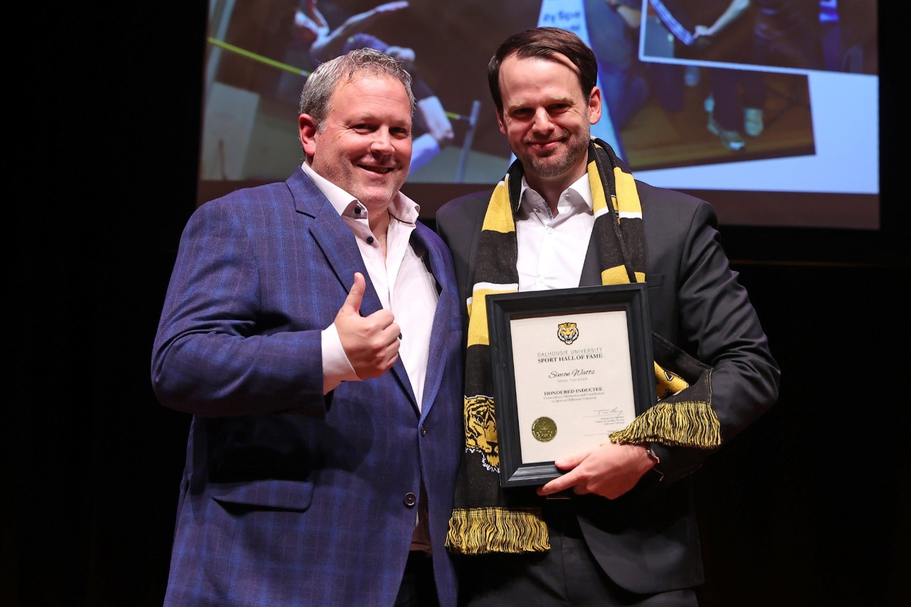 A man wearing a black and gold scarf holds a framed certificate standing next to a man giving a thumbs up on stage at an event.