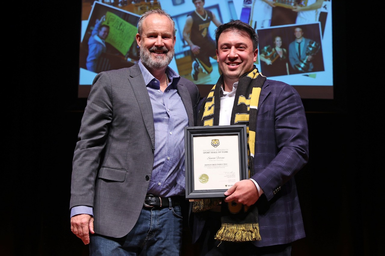 A man wearing a black and gold scarf holds a framed certificate standing next to a man smiling on stage at an event.