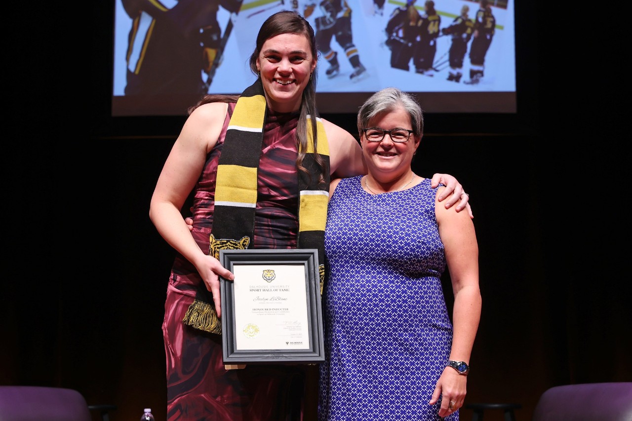 A woman wearing a black and gold scarf holds a framed certificate standing with her arm around a woman smiling on stage at an event.