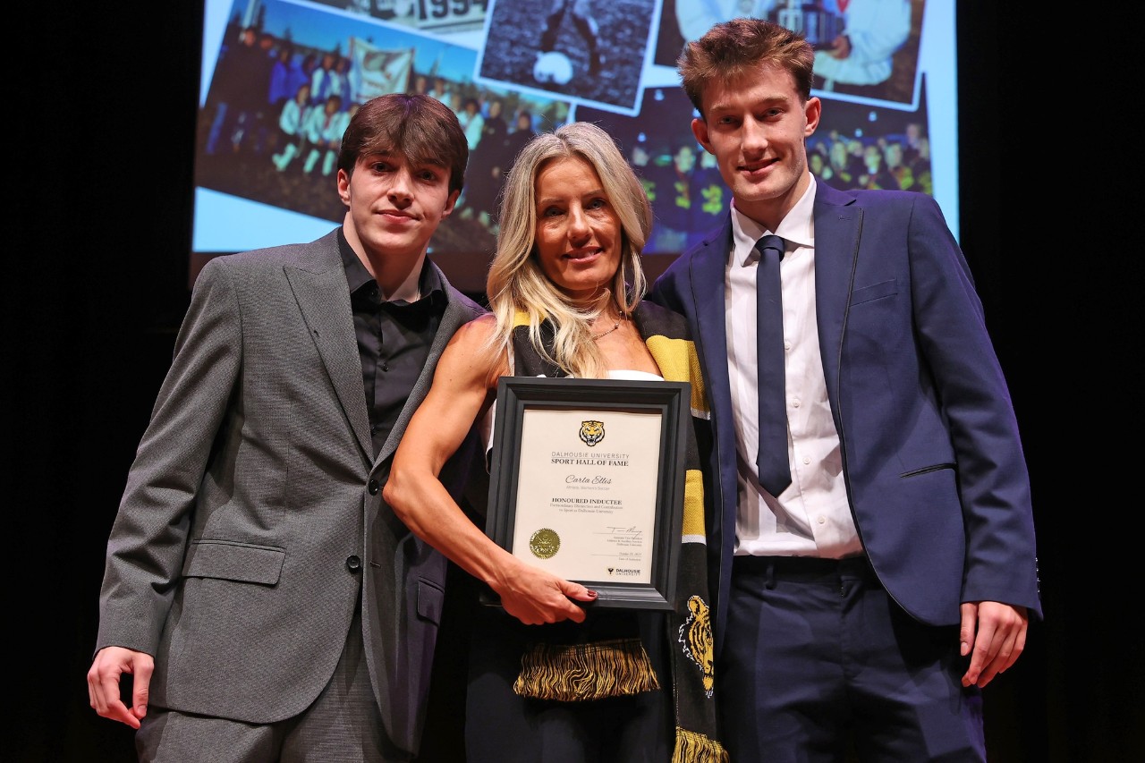 A woman wearing a black and gold scarf holds a framed certificate standing between two young men on stage at an event.