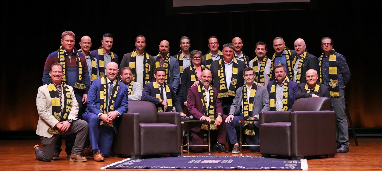 A group of former student-athletes wearing black and gold scarves pose on stage at an event.