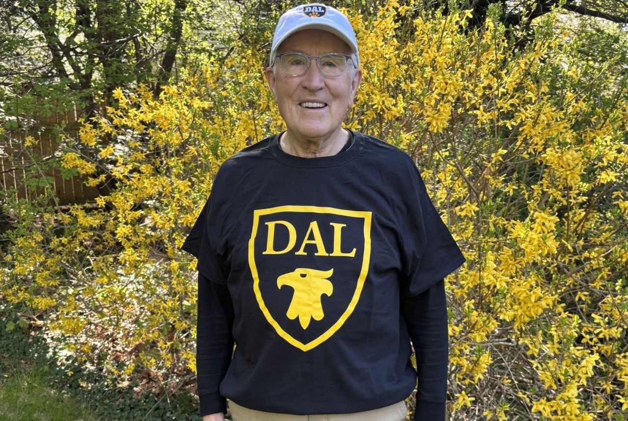 Alan Hayman is wearing a cap, glasses, and a Dal shirt while staning in his garden