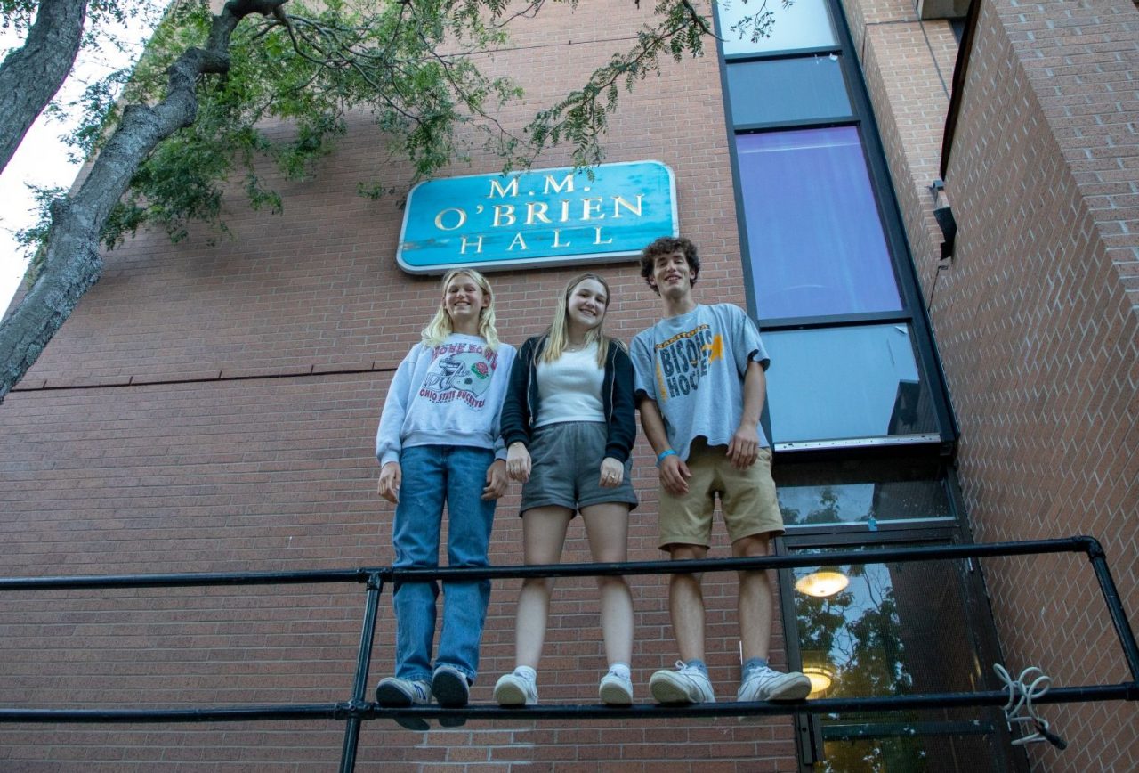 Three students stand on the railing outside a tall brick residence building dressed for warm weather and a leafy tree over top.