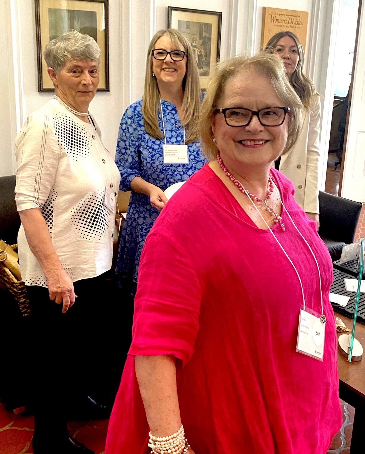Cynthia Pilichos looks beyond the camera, smiling while three other women smile in the background at the reception area of an event.