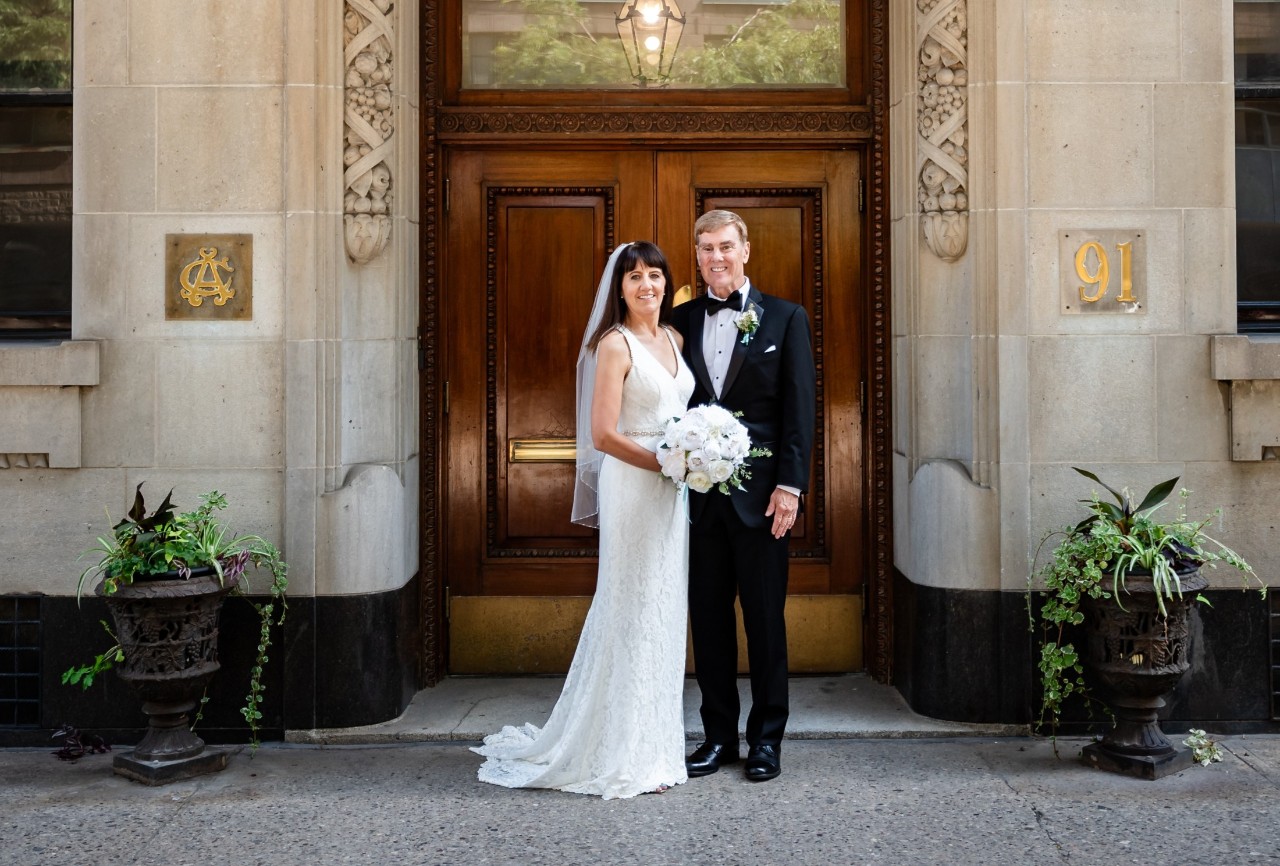 A man and a woman stand together in wedding attire with the bride holding a bouquet of white roses outside elaborate wooden double doors of a stone building.