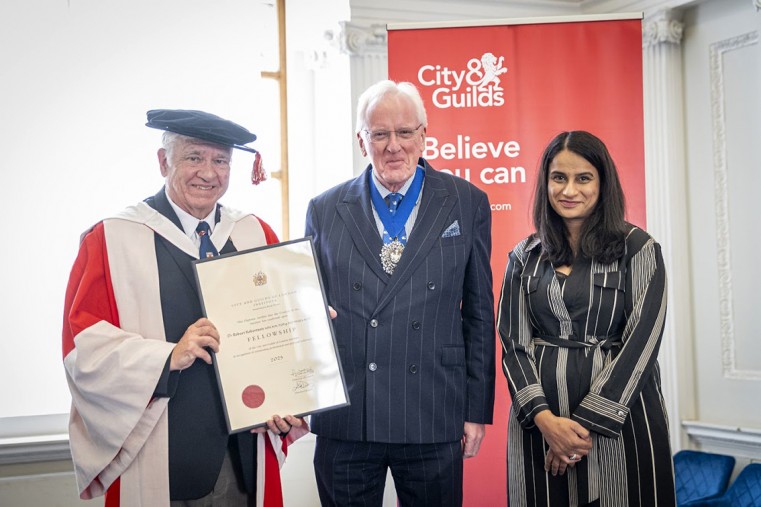 A man in convocation cap and gown holds a framed certificate standing alongside another man and woman.