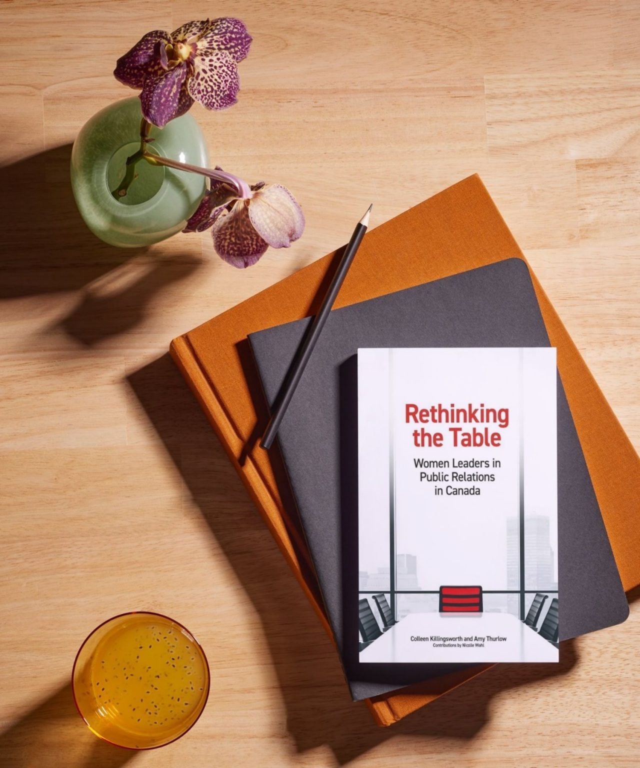 Book cover set upon stack of 2 notebooks with a pencil and flower in a vase.