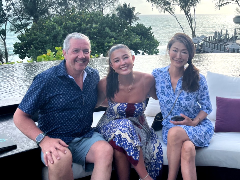 Gibbons sits with his daughter and spouse on an outdoor couch at a resort overlooking the water.