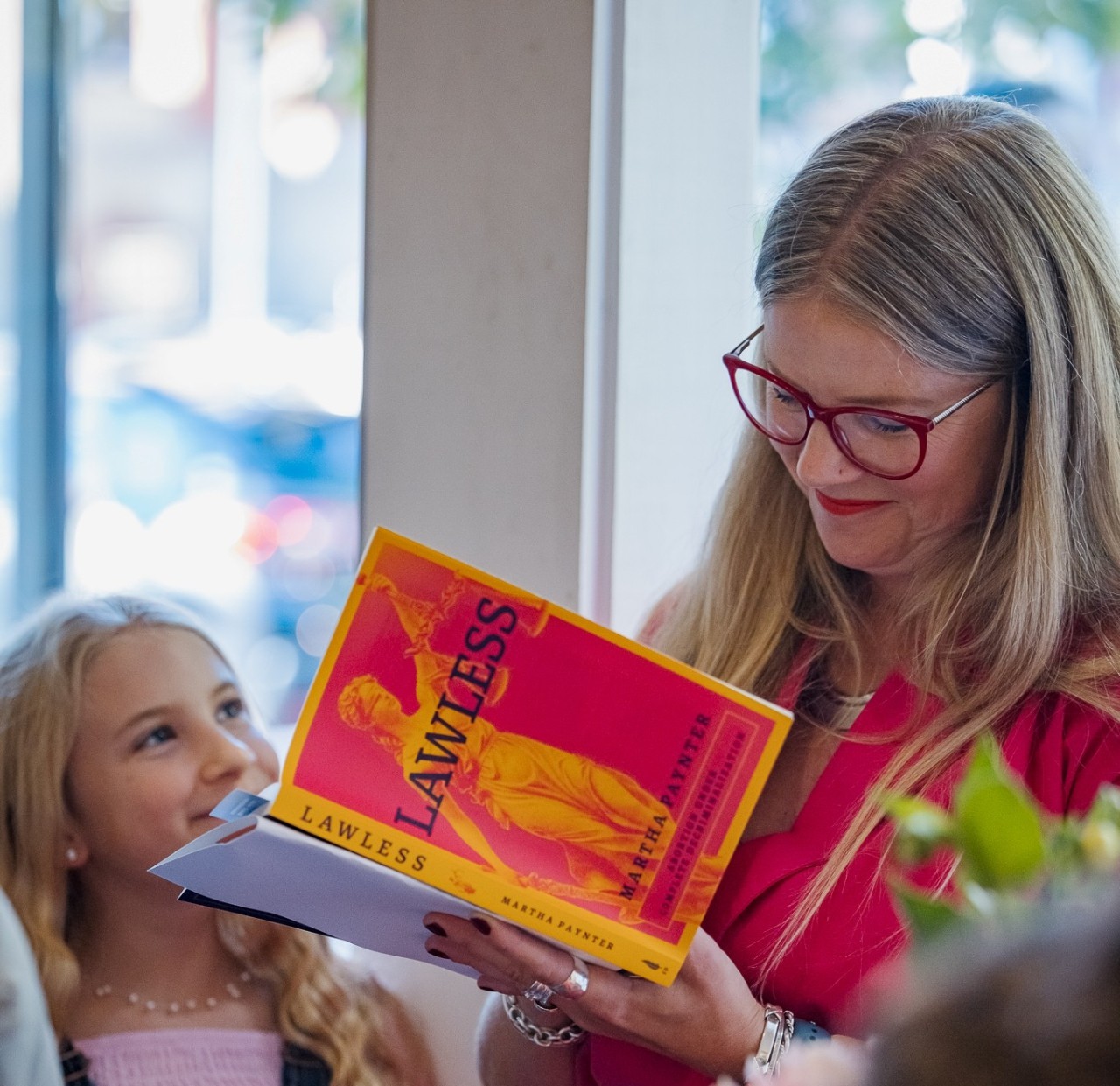 A woman wearing glasses smiles while signing a book as a younger person looks up at her.