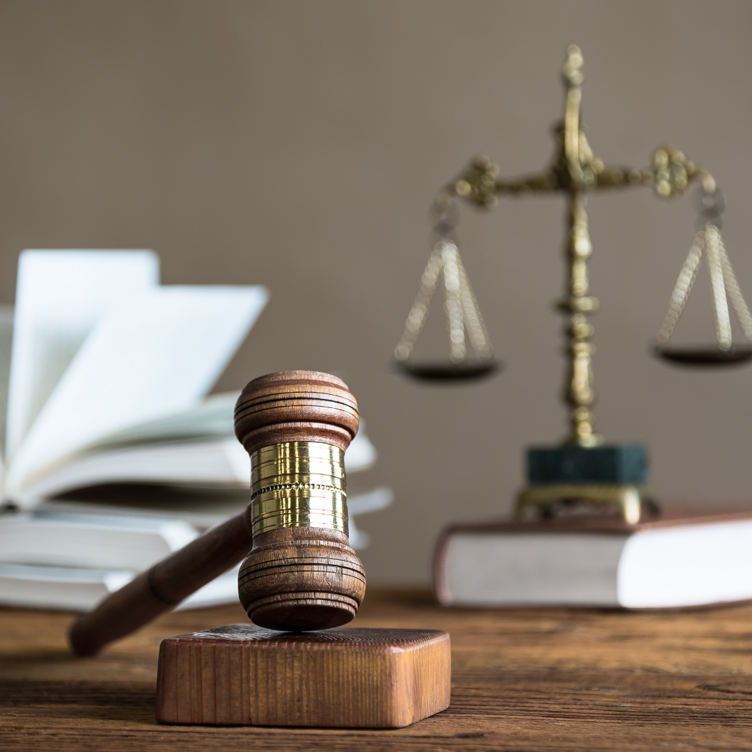 A gavel rests on a block on a desk with balance scales and books in the background.