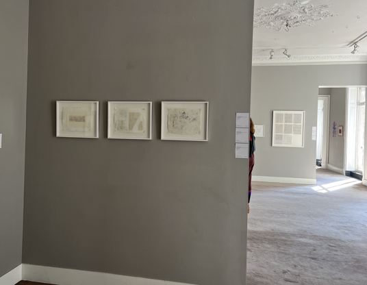 Three framed works of art hanging in a row on an interior grey wall, opening into another room.