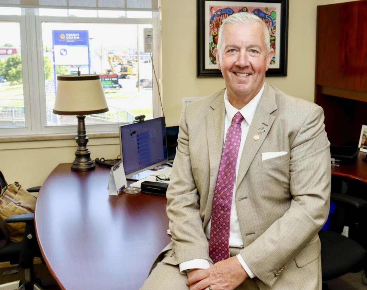 Gibbons dressed in a light-coloured suit and tie perches casually on the edge of a desk in an office, smiling with hands clasped.
