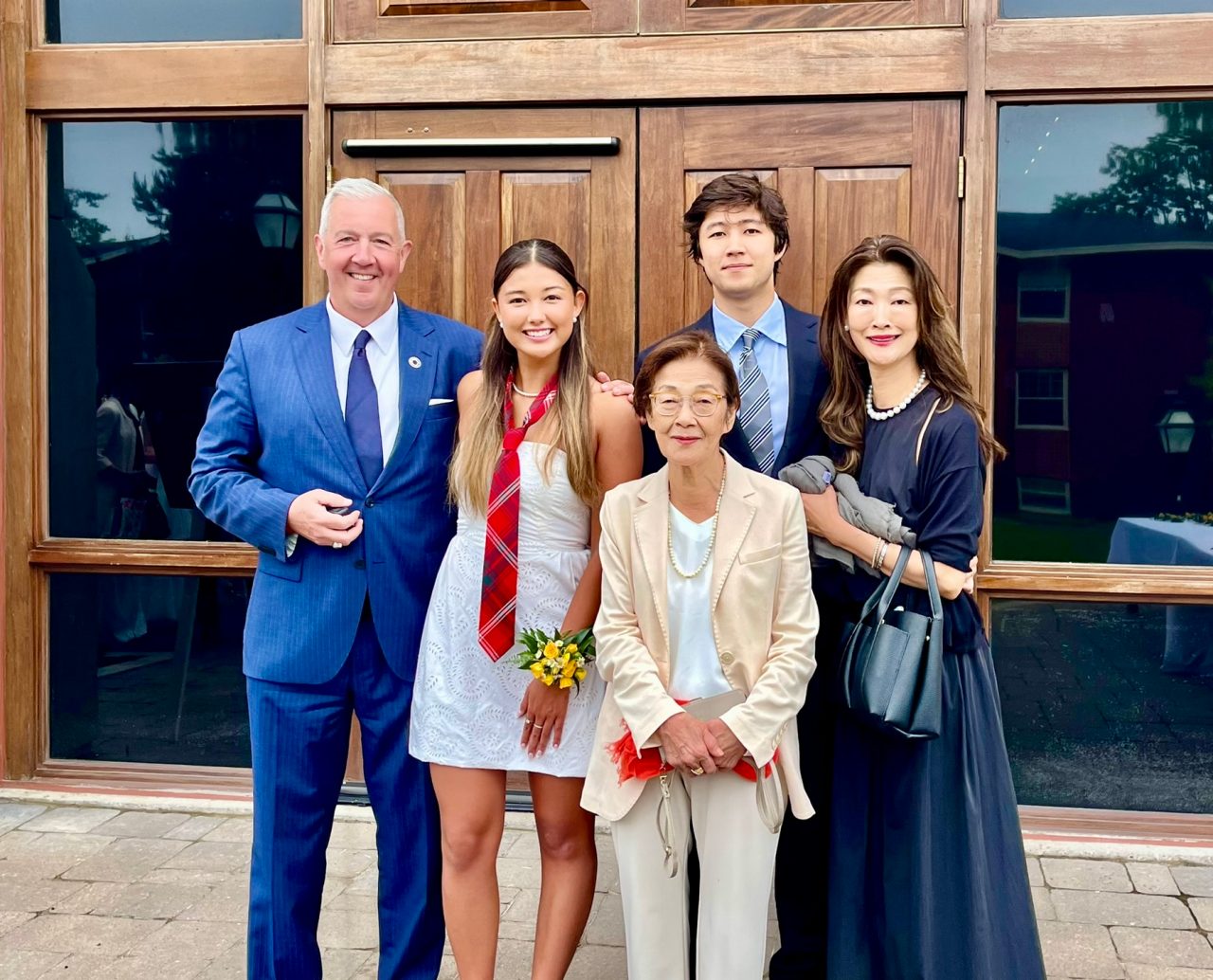 Family members stand outdoors in front of large wooden doors and glass panes, dressed formally for an event.
