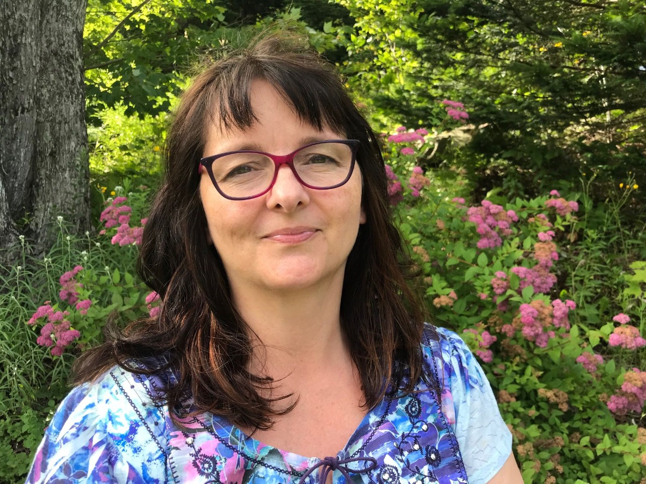 A woman wearing glasses and smiling slightly outdoors with trees, flowers and foliage in the background.