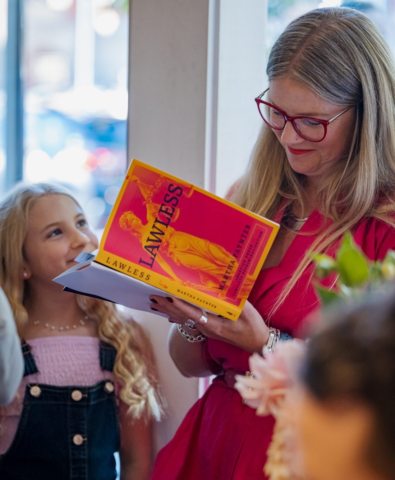 A woman wearing glasses smiles while signing a book as a younger person looks up at her.