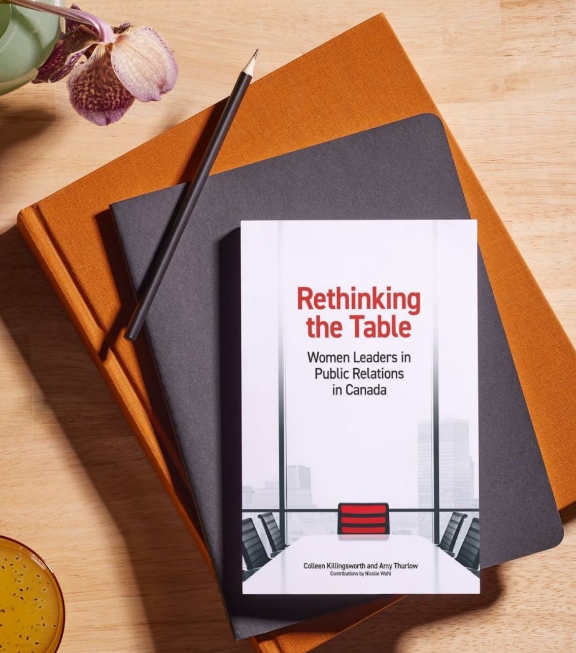 Book cover set upon stack of 2 notebooks with a pencil and flower in a vase.