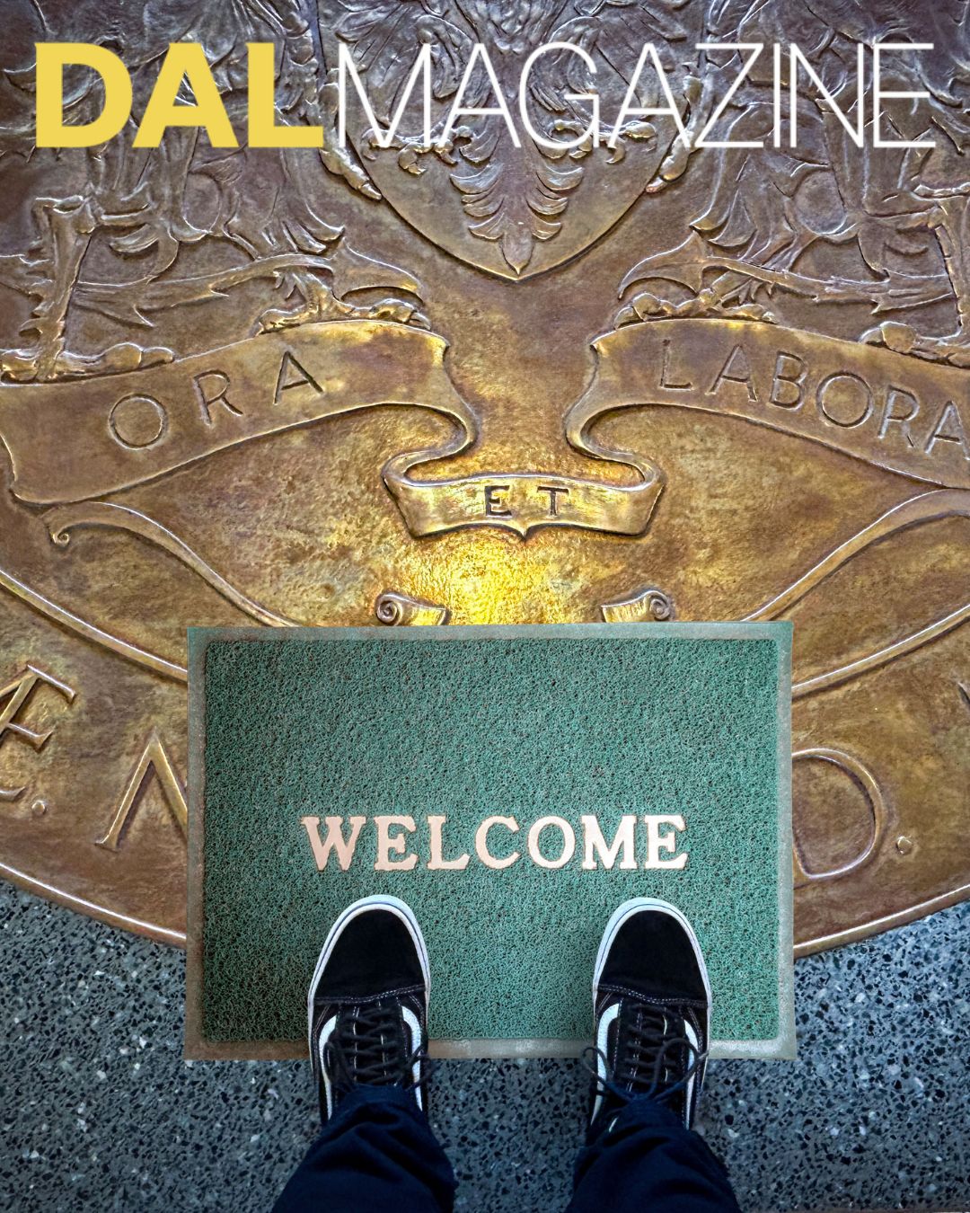A person's feet in black and white sneakers with jeans showing stand on a green welcome mat atop the Dalhousie University Coat of Arms engraved on the lobby floor. 