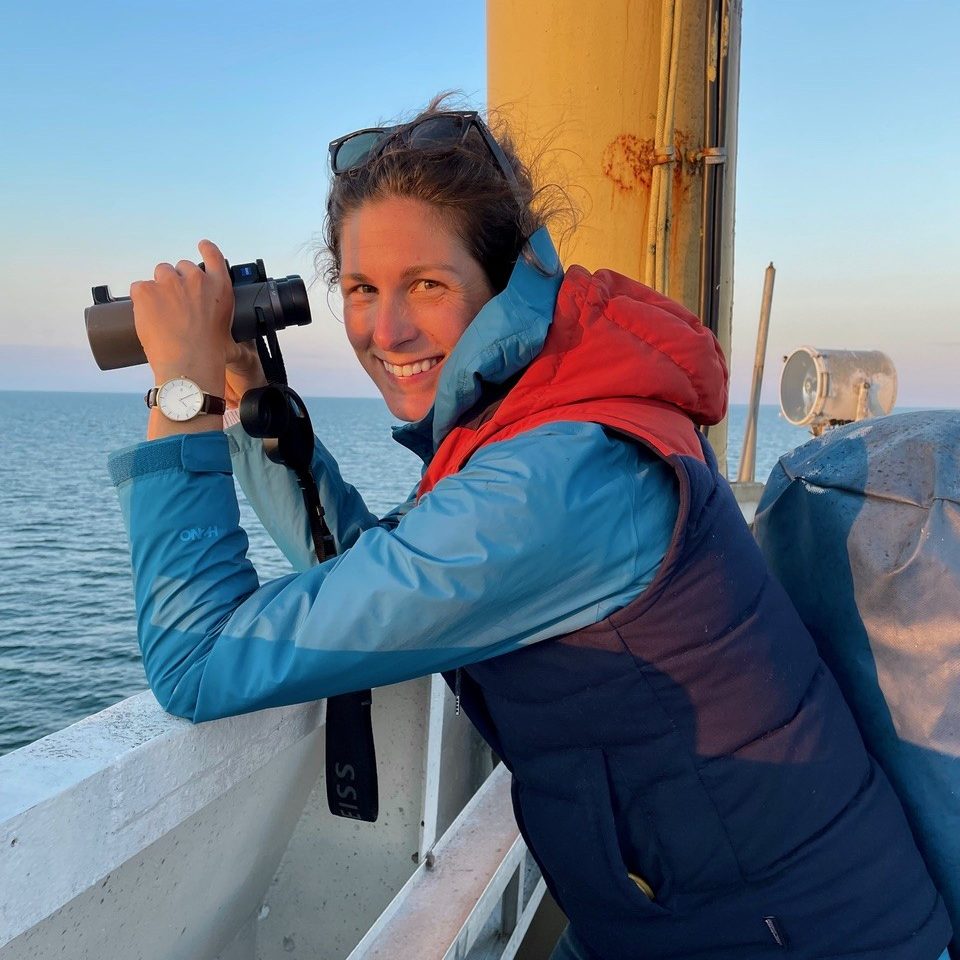 A woman smiling aboard a boat wearing a rain jacket and warm vest holding binoculars to look out to sea.