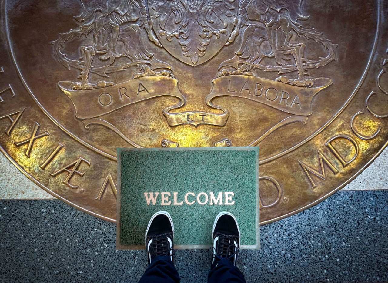 A person's feet in black and white sneakers with jeans showing stand on a green welcome mat atop the Dalhousie University Coat of Arms engraved on the lobby floor. 