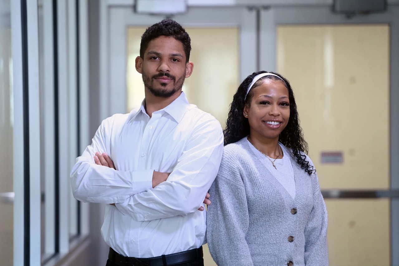 James Dixon stands with arms crossed in a white dress shirt standing slightly turned back to back with Portia Wright smiling in a grey button-up sweater.