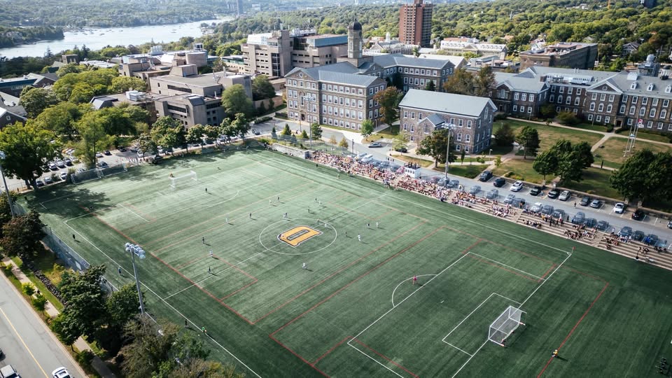 An aerial view of Wickwire Field at Dalhousie showing the surrounding campus buildings, trees, parking and distant waterway.