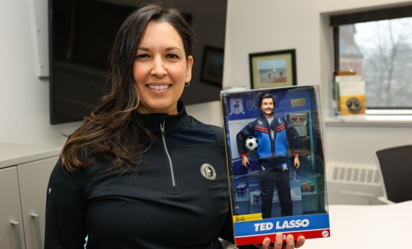A woman wearing an athletic top smiles holding a doll of Ted Lasso in its packaging.