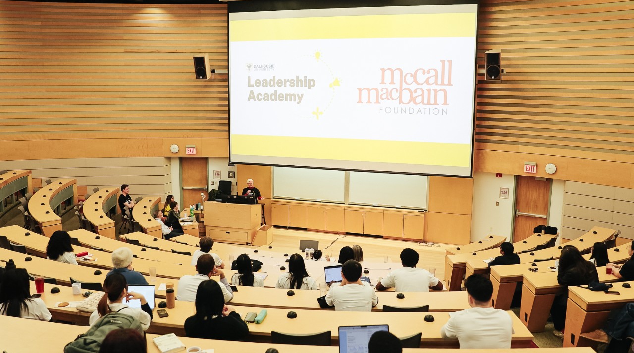 Students are sitting in a U-shaped auditorium with a projector screen reading 'McCall MacBain Leadership Academy' at the front.