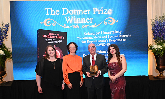 A man in a suit holds an award styled like a book standing in a row with three woman against a screen at a semi-formal event. 