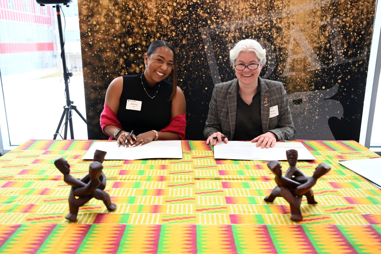 Hill and Dr. Brooks smiling while reviewing papers and seated a table adorned with a colourful table cloth and culturally-significant wooden carvings.