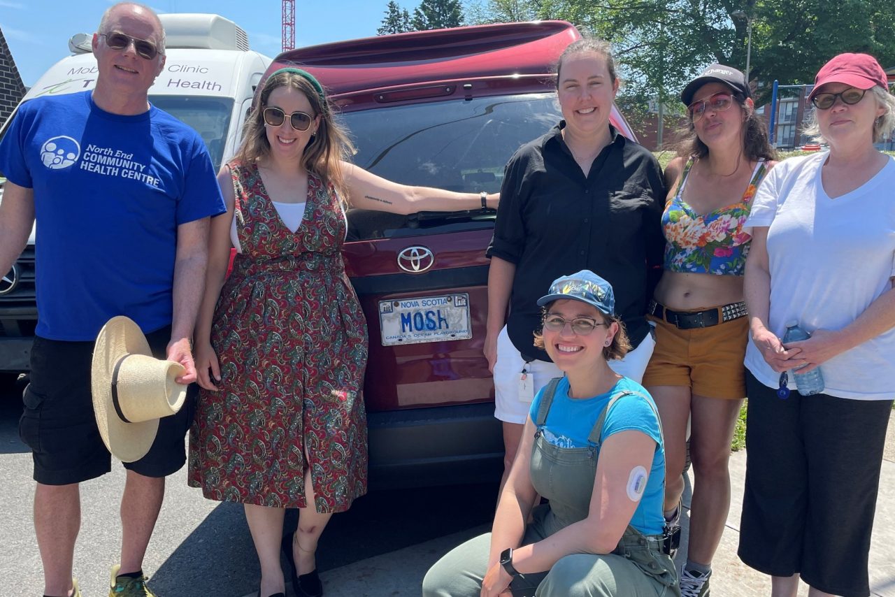 A woman in overalls, t-shirt and ball cap crouches down in front of five other people standing at the back of a red van in fine summer weather.