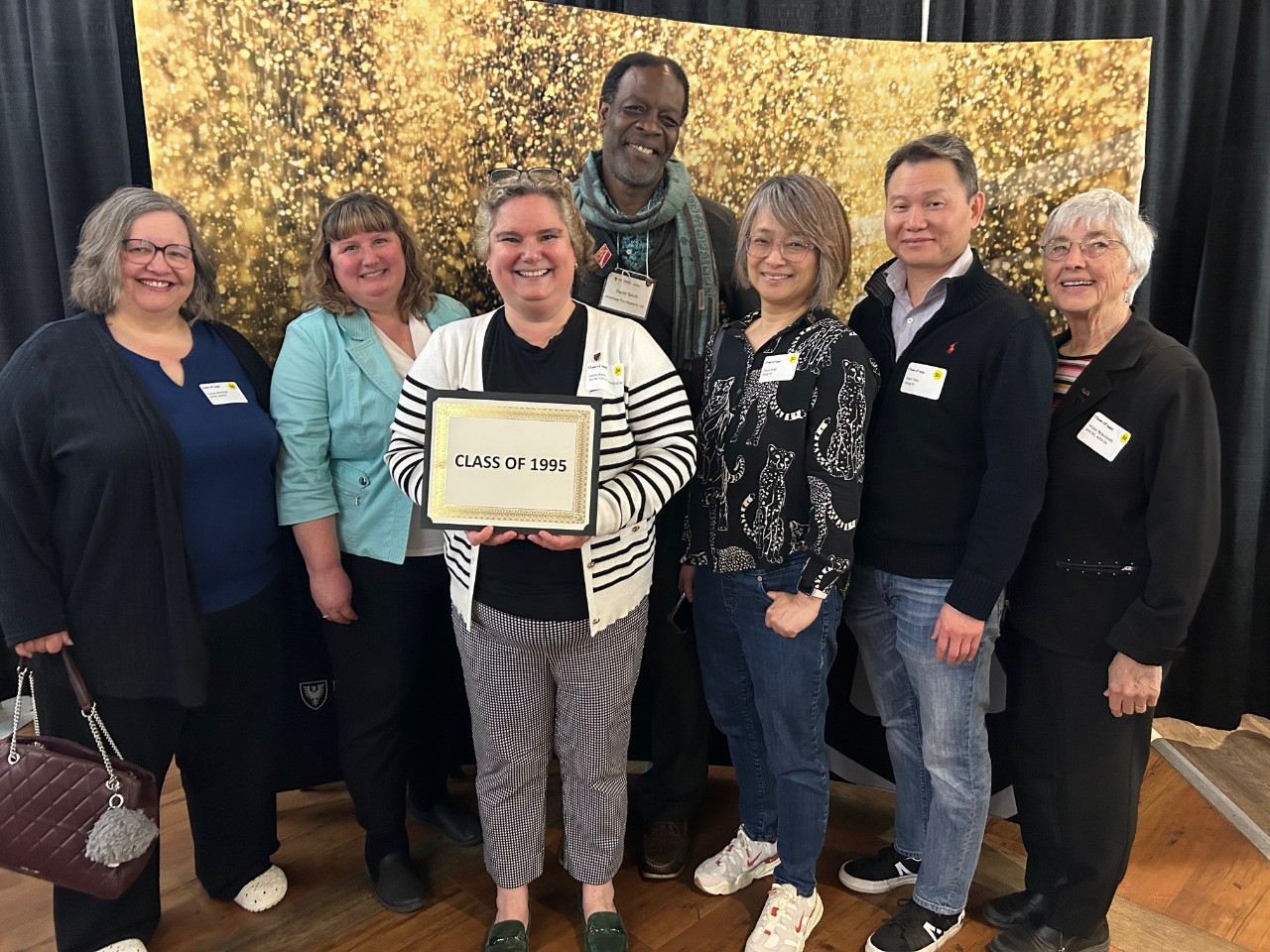 A group of 7 people pose together holding a sign "Class of 1995" against a shiny gold backdrop.