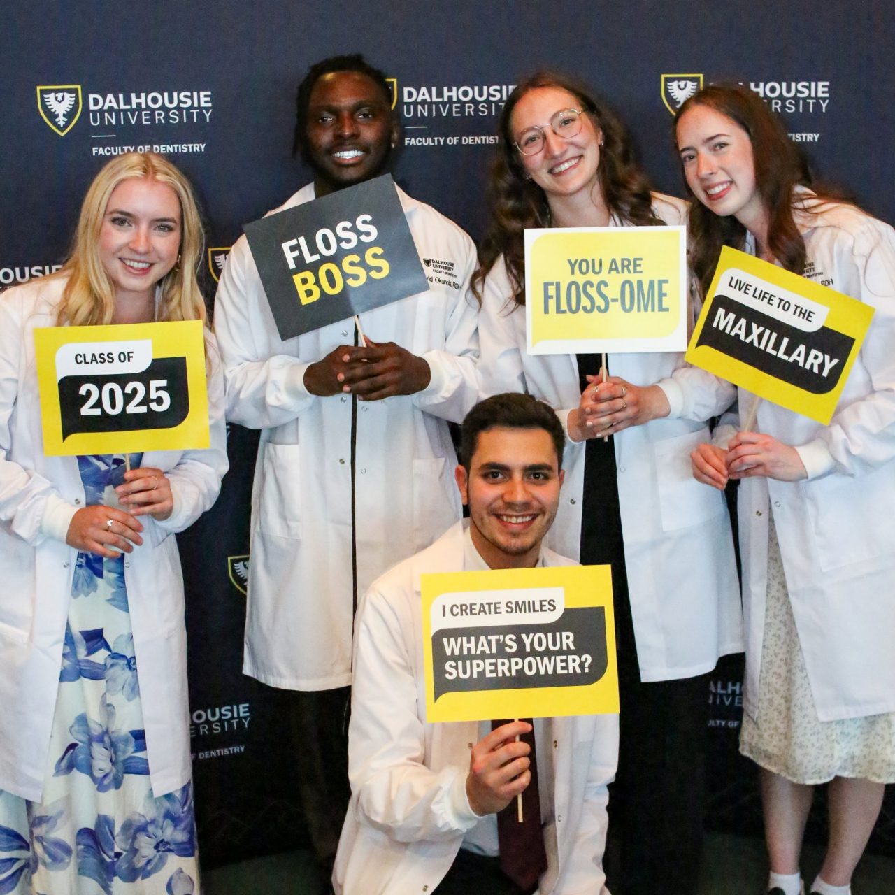 Five people holding signs wearing white lab coats over semi-formal attire at a celebratory event.