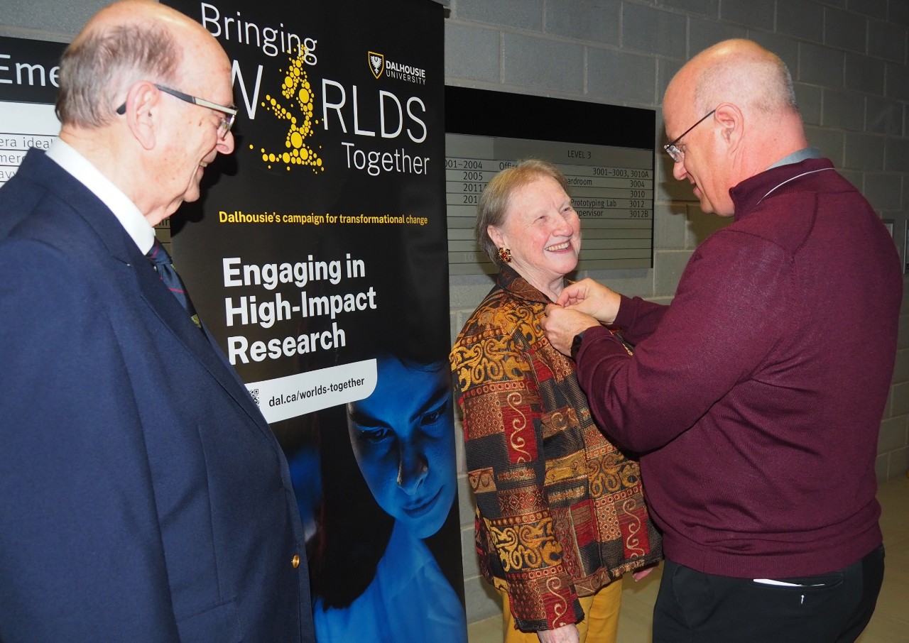 Gilkie looks on as Barker's pin is attached to her jacket lapel, both smiling, at the event. 