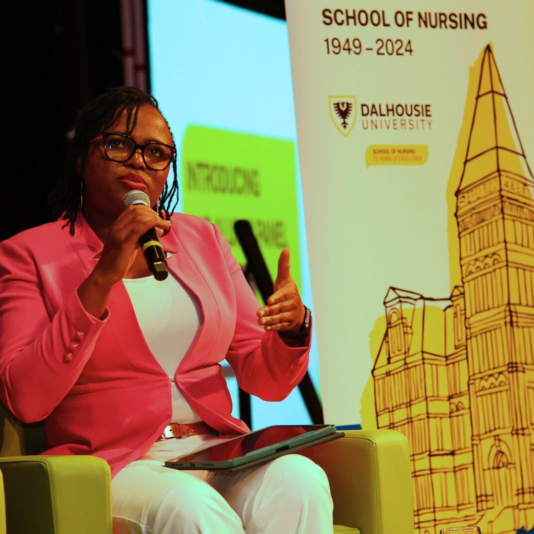A woman is seated in a yellow sofa chair wearing a pink blazer and speaking into a microphone on stage at an event.