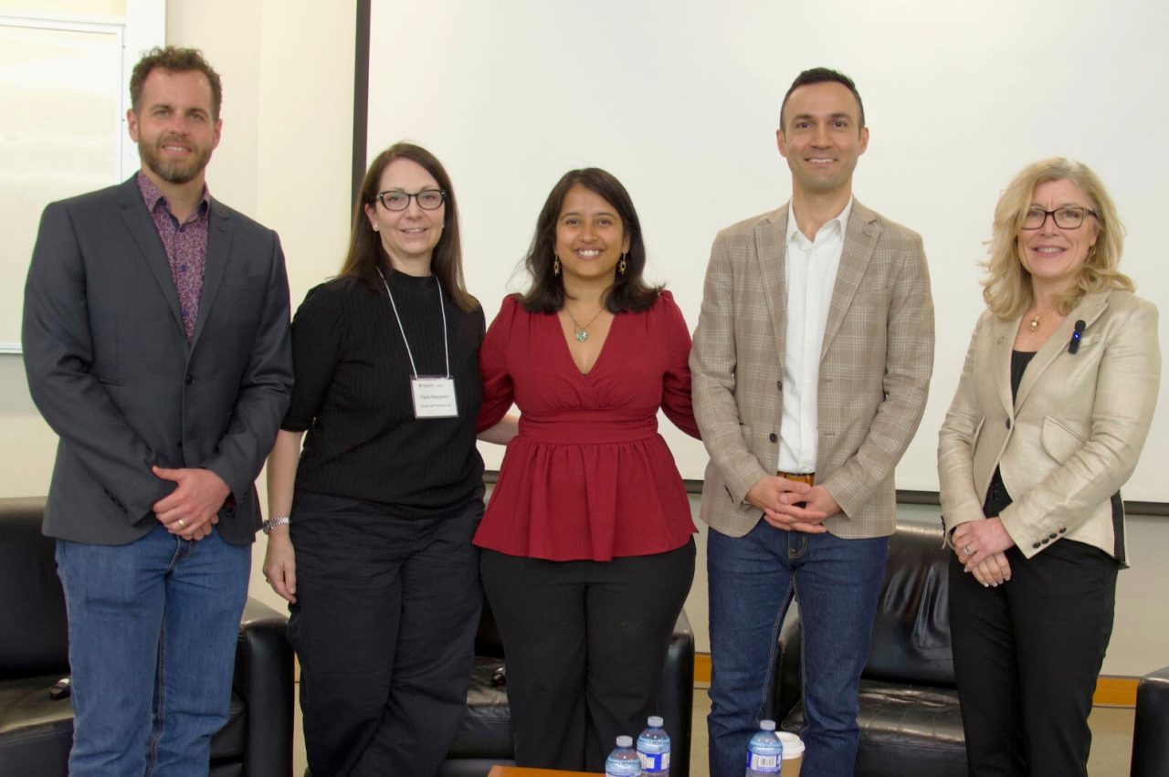 Five event panelists stand in front of soft seating smiling with hands clasped in front.
