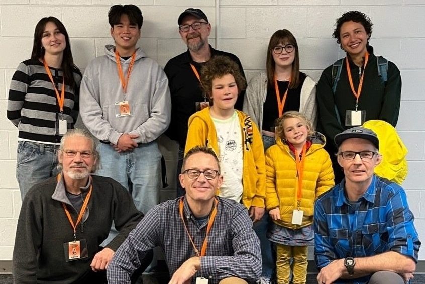 A group of ten people, including adults and children, pose for a photo indoors against a white wall, all wearing orange lanyards.