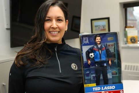A woman wearing an athletic top smiles holding a doll of Ted Lasso in its packaging.