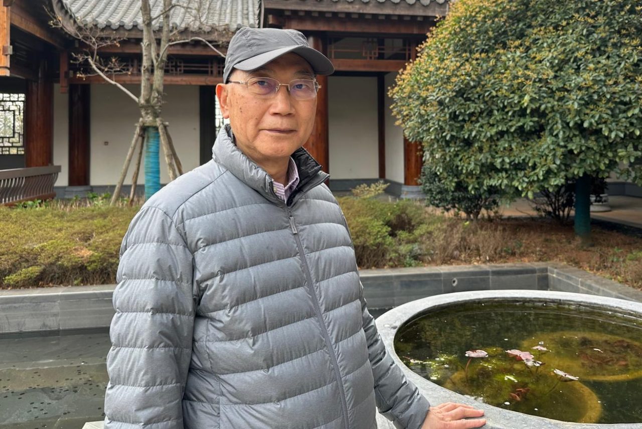 Peter Chow wearing a grey puffy jacket, grey ball cap and glasses smiling slighting next to a small pond feature outside a building with sloping roofs.