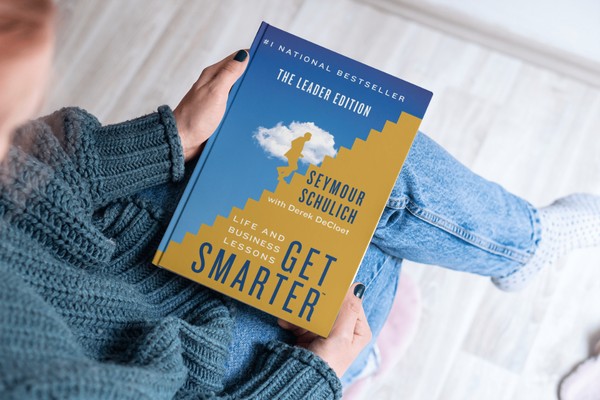A person in jeans and a sweater holding the book Get Smarter: Life And Business Lessons.