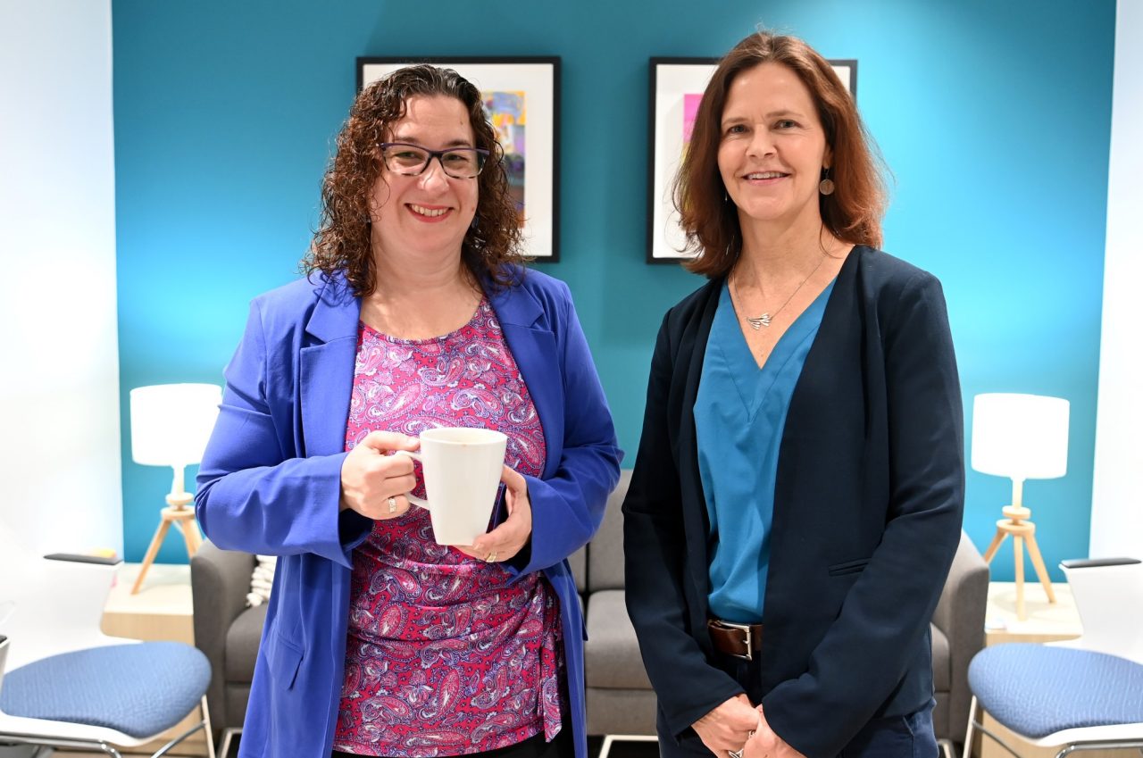 Two women stand together smiling, one holds a mug, in a brightly decorated office space.