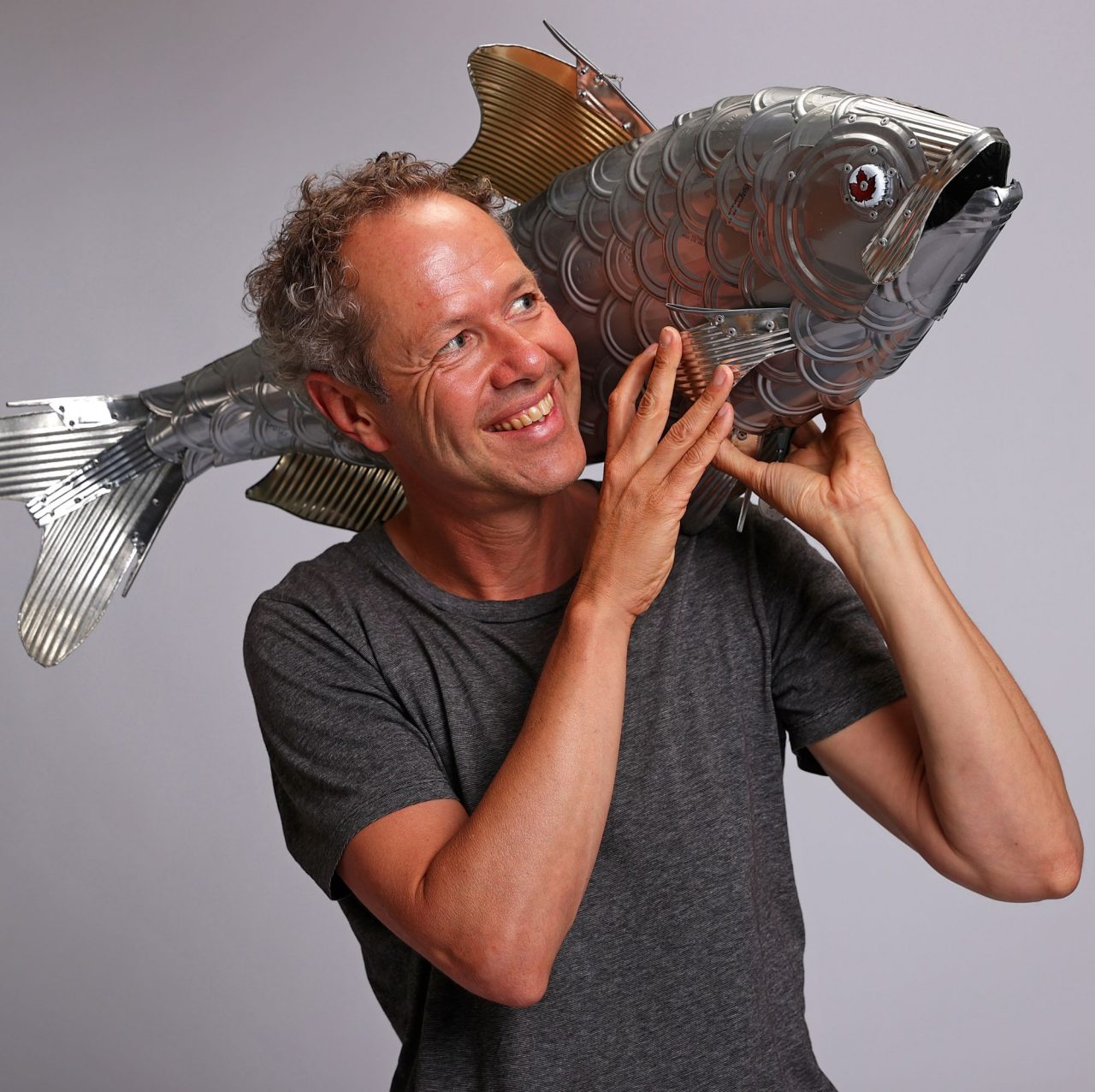 Smiling man holding a large metallic fish sculpture on his shoulder, wearing a grey t-shirt against a plain background.