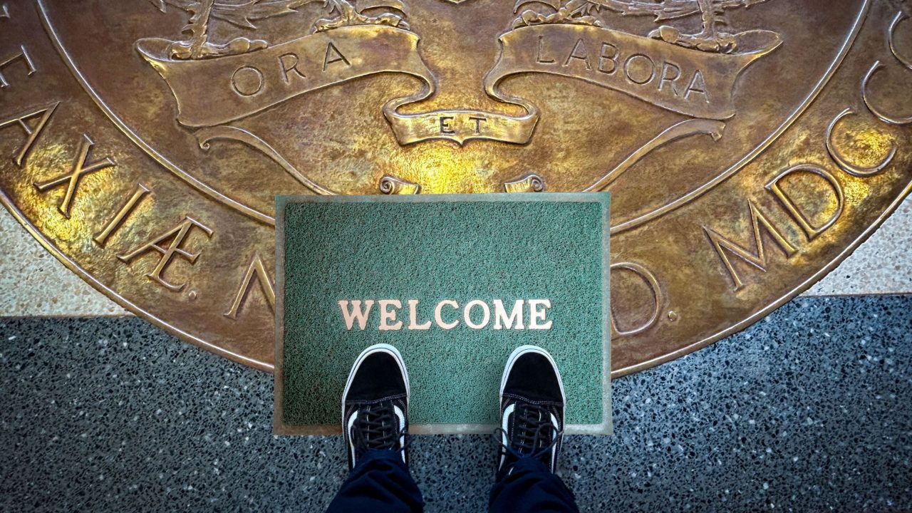 A person's feet in black and white sneakers with jeans showing stand on a green welcome mat atop the Dalhousie University Coat of Arms engraved on the lobby floor. 