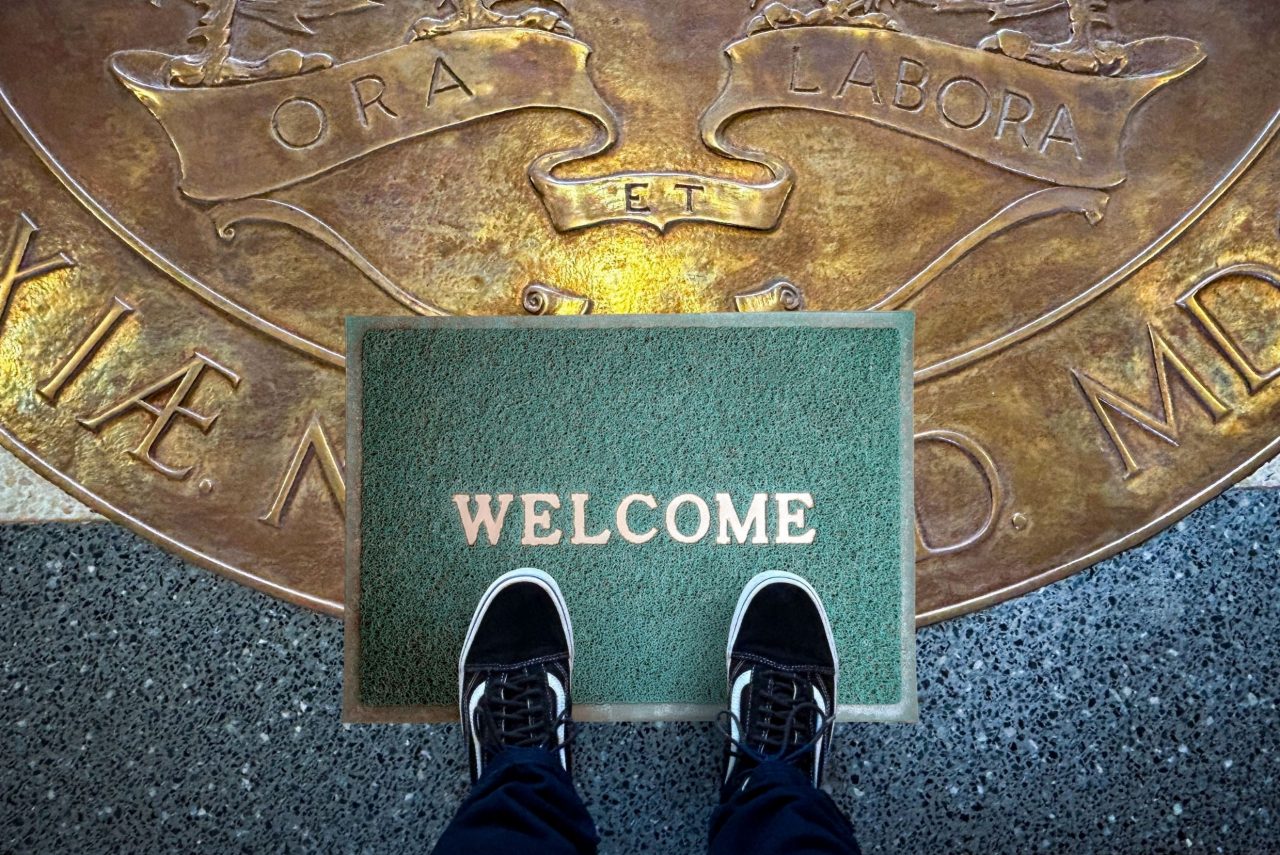 A person's feet in black and white sneakers with jeans showing stand on a green welcome mat atop the Dalhousie University Coat of Arms engraved on the lobby floor. 