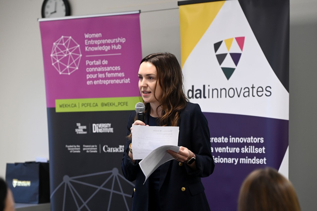 A woman speaks into a microphone while holding notes at a conference with banners in the background.