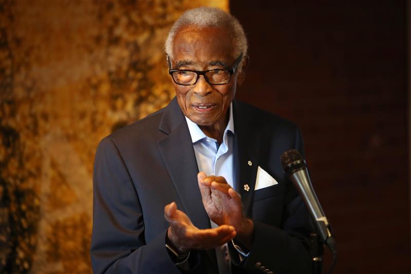 Donald Oliver on stage at a formal event standing next to a microphone on a stand, clapping his hands.