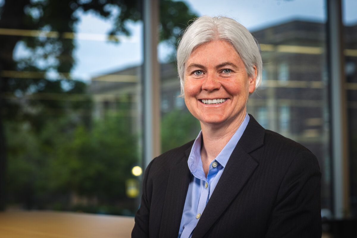 A woman in a suit in front of a window with buildings and trees visible in the background