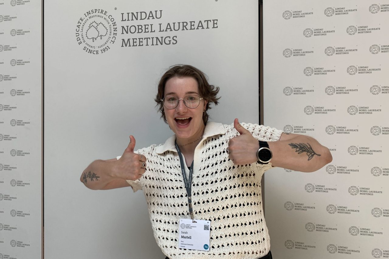 Sarah Martell standing in front of event signage wearing a nametag on a lanyard and giving two thumbs up.