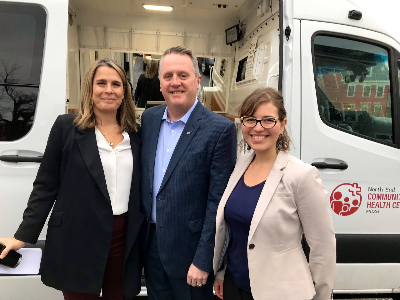 Two women and a man dressed in business casual attire smile alongside a large van with the sliding door open.