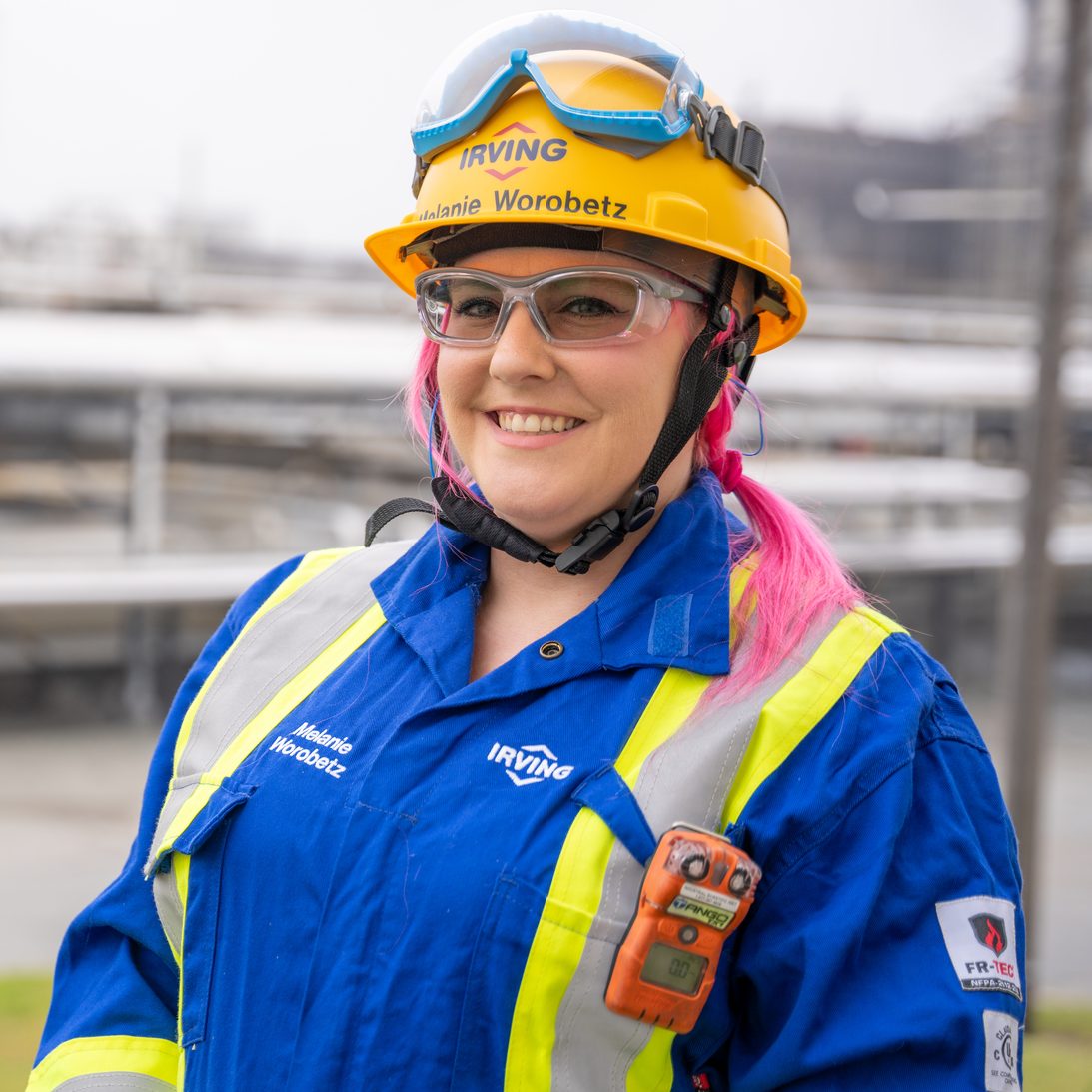 A female-presenting person with pink hair wearing safety glasses, a hard hat and coveralls, smiling on a work site.