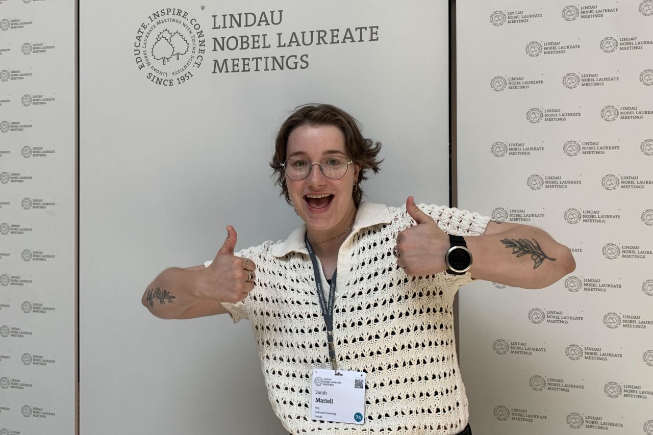 Sarah Martell standing in front of event signage wearing a nametag on a lanyard and giving two thumbs up.